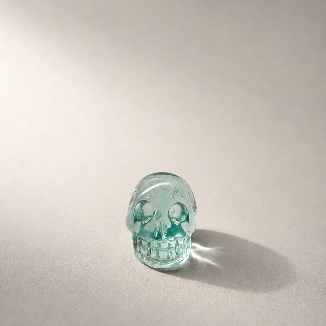 A small translucent blue obsidian skull figurine standing on a neutral gray surface with a soft shadow cast to the right.