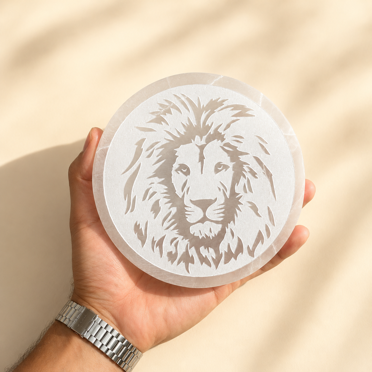 Hand holding a round selenite charging plate with a lion design against a beige background