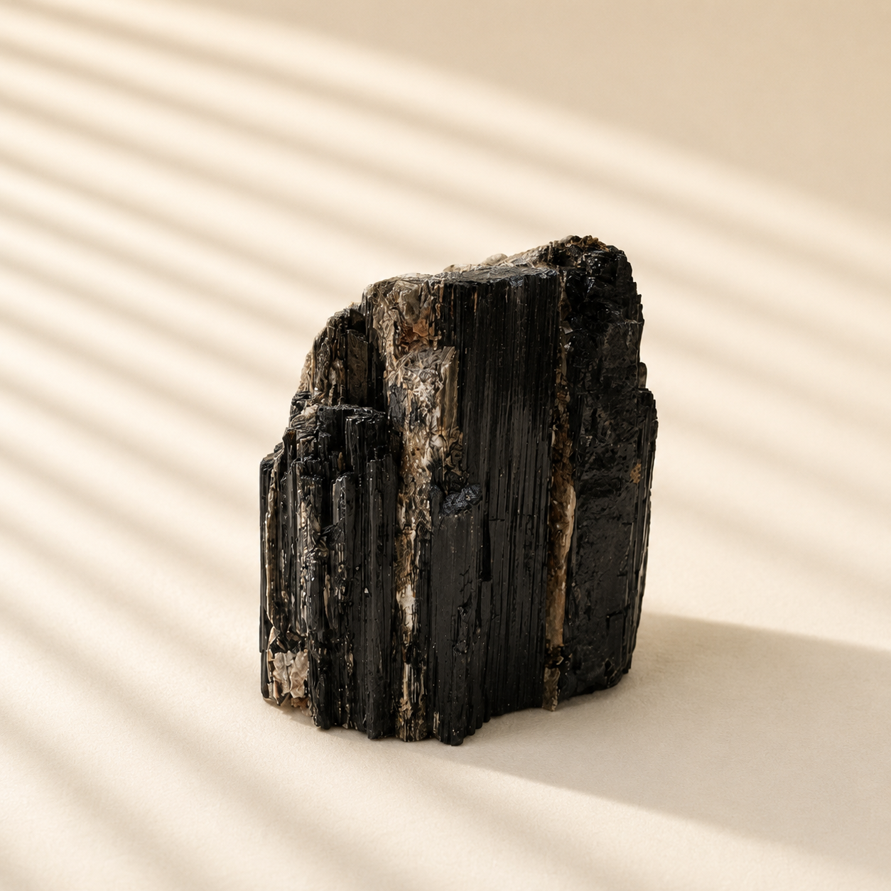 A rough black tourmaline mineral specimen with vertical striated crystal structure, standing upright on a warm beige surface under soft diagonal shadows.