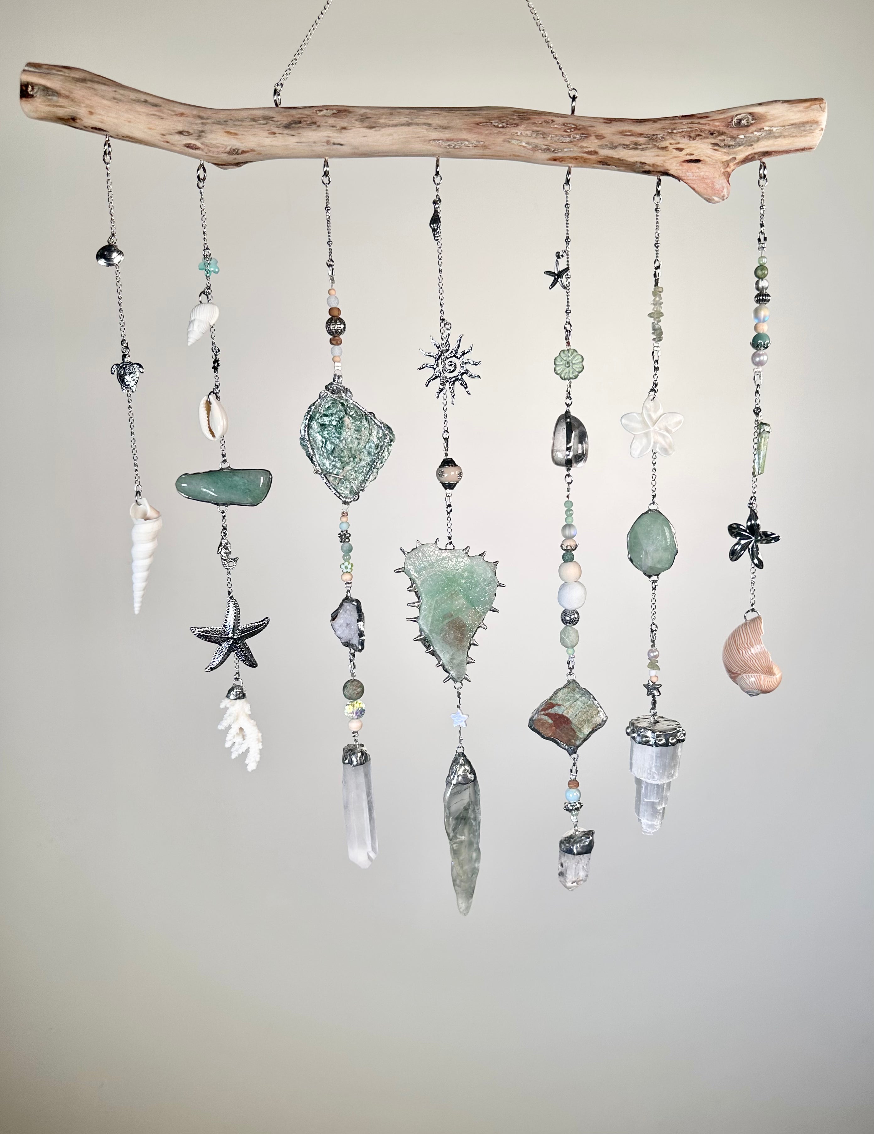 Mermaid themed crystals hanging from a natural driftwood branch. The crystal wall hang features various seashells, starfish, crystals, and small beads in muted sage green, gray, white, and soft pink tones, suspended against a plain light gray background.