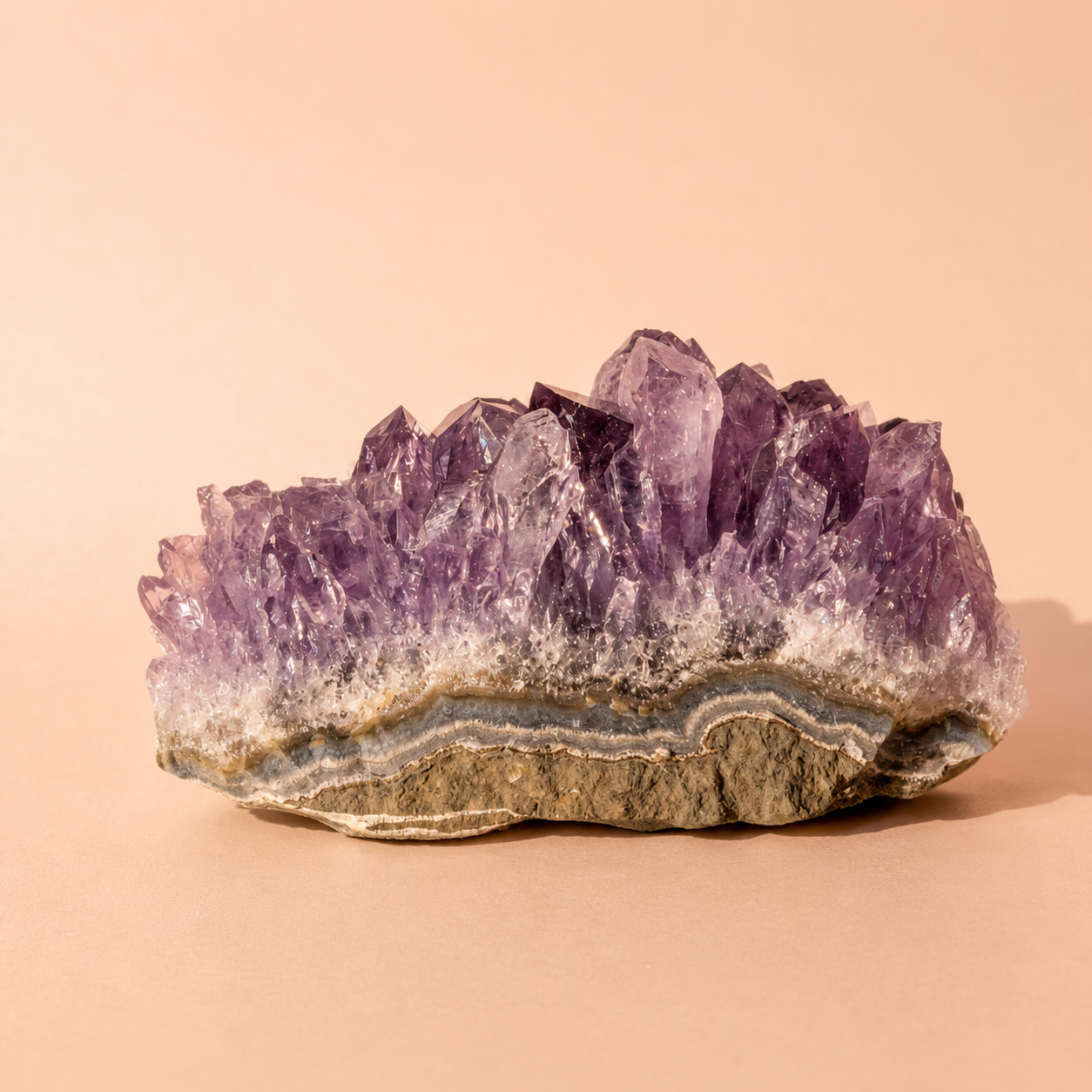 A natural amethyst crystal cluster geode specimen with purple crystalline points rising from a gray and beige banded matrix, photographed on a warm peach studio background.