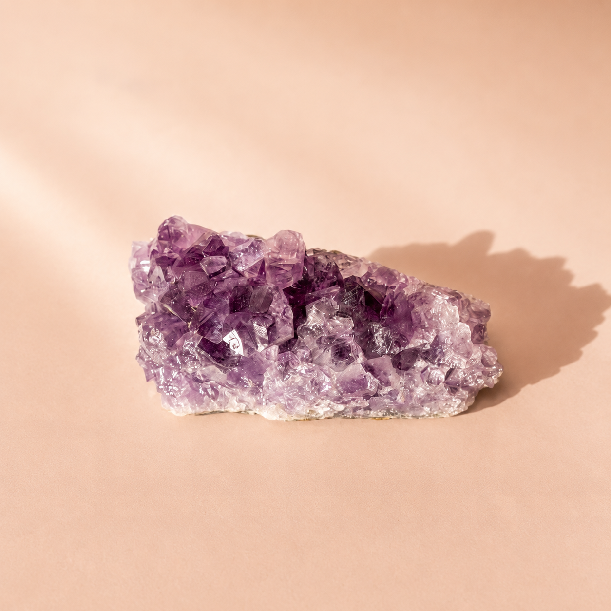 A raw amethyst crystal cluster with prominent purple and lavender crystalline formations on a light beige background, casting a soft shadow to the right.