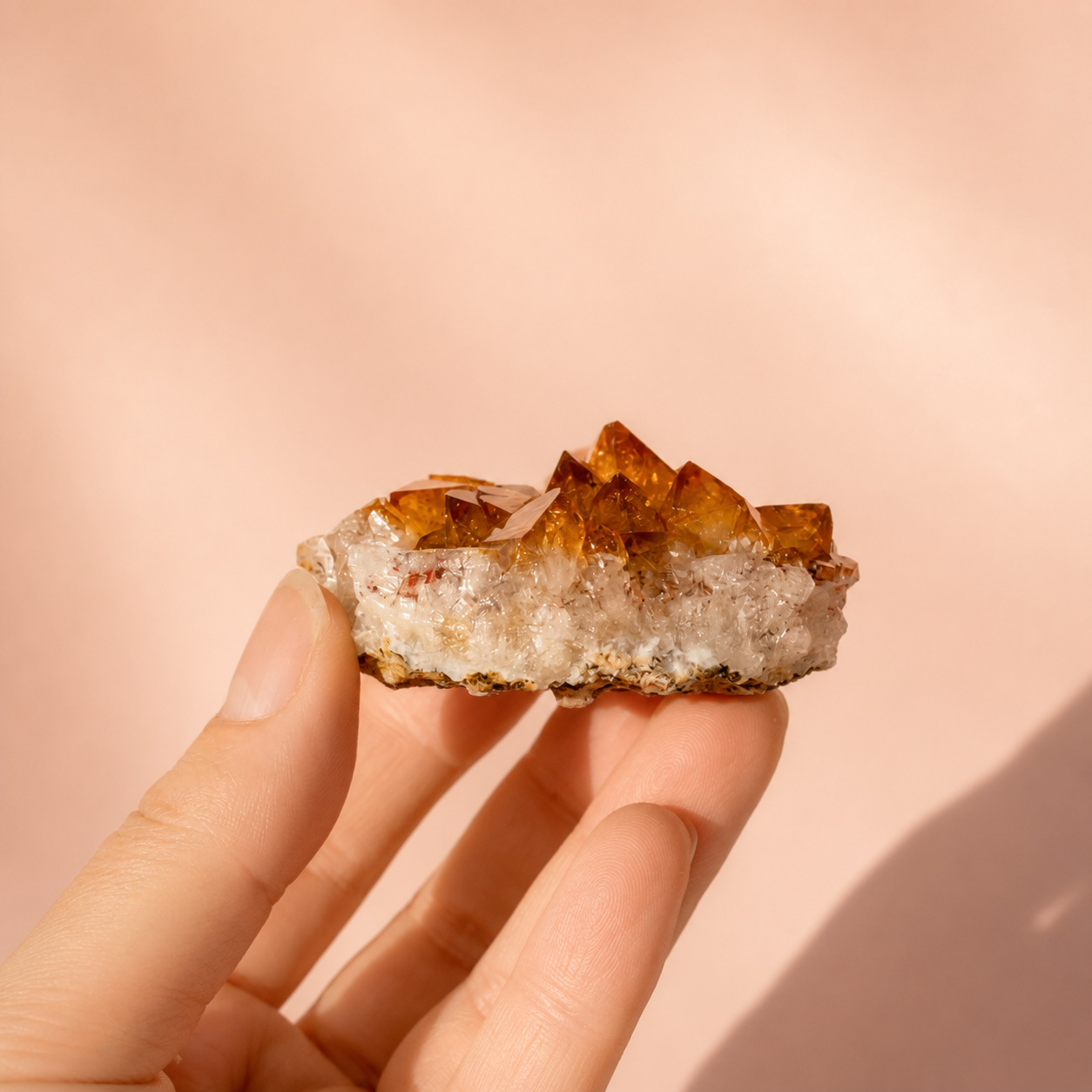 A hand holding a small citrine crystal cluster with amber-brown pointed tips and a pale frosted base against a soft peach-pink background.