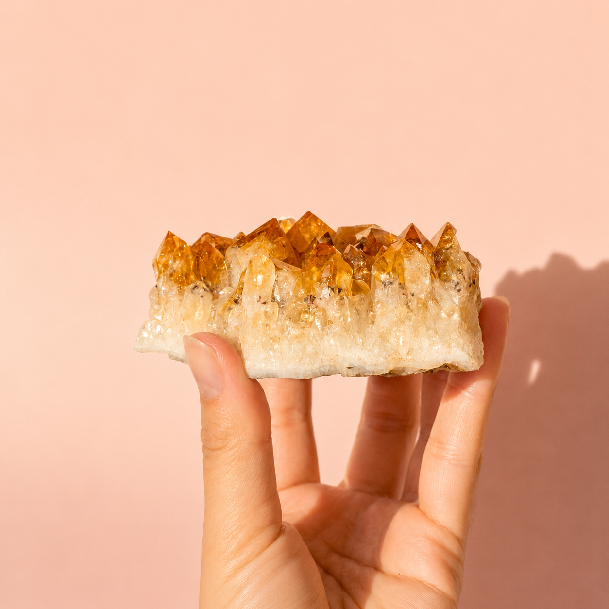 A hand holding a natural crystal cluster with amber-orange and translucent white points against a soft peach-pink background.