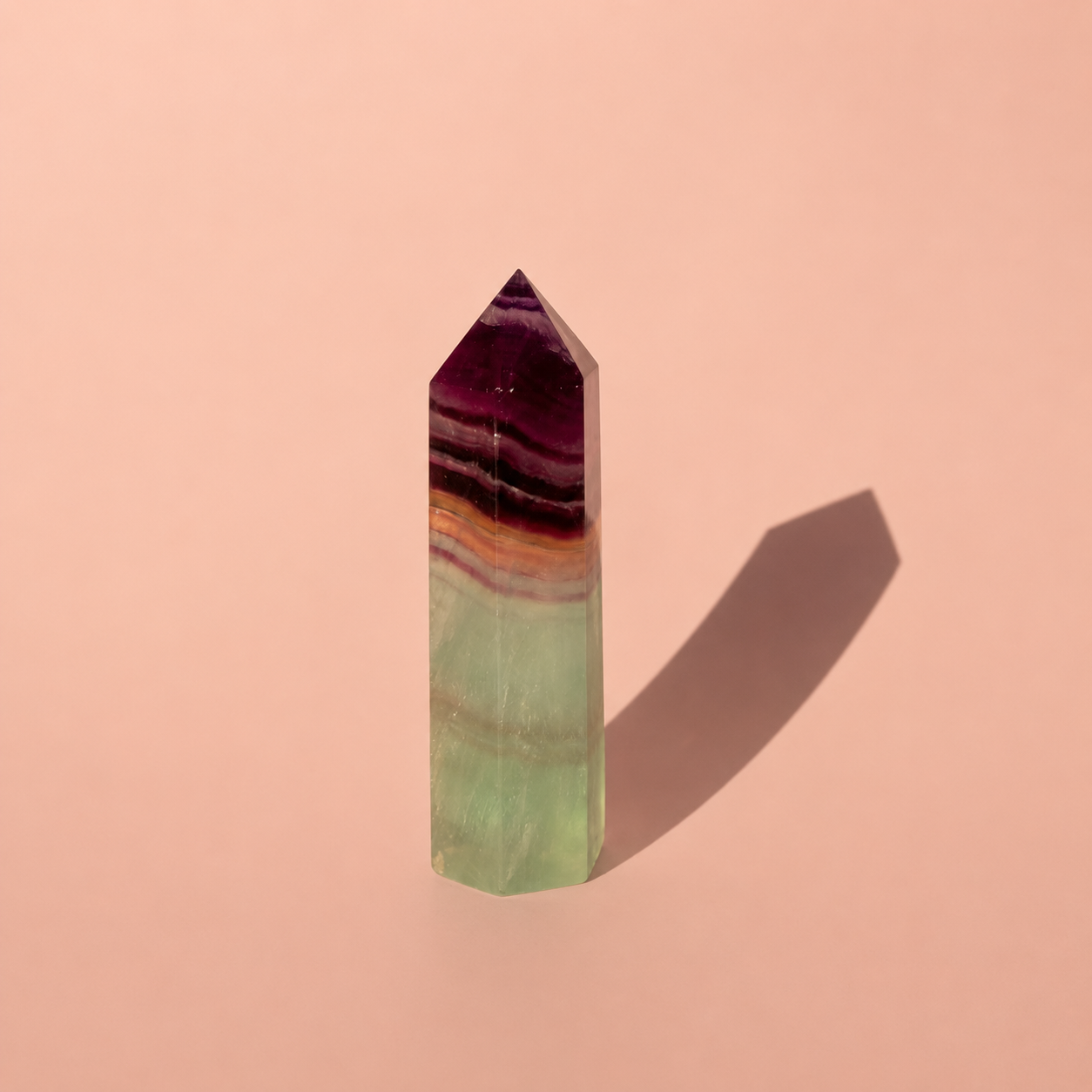 A polished fluorite crystal point with horizontal banding of purple, orange, and light green, standing upright on a solid pink background with a soft shadow.