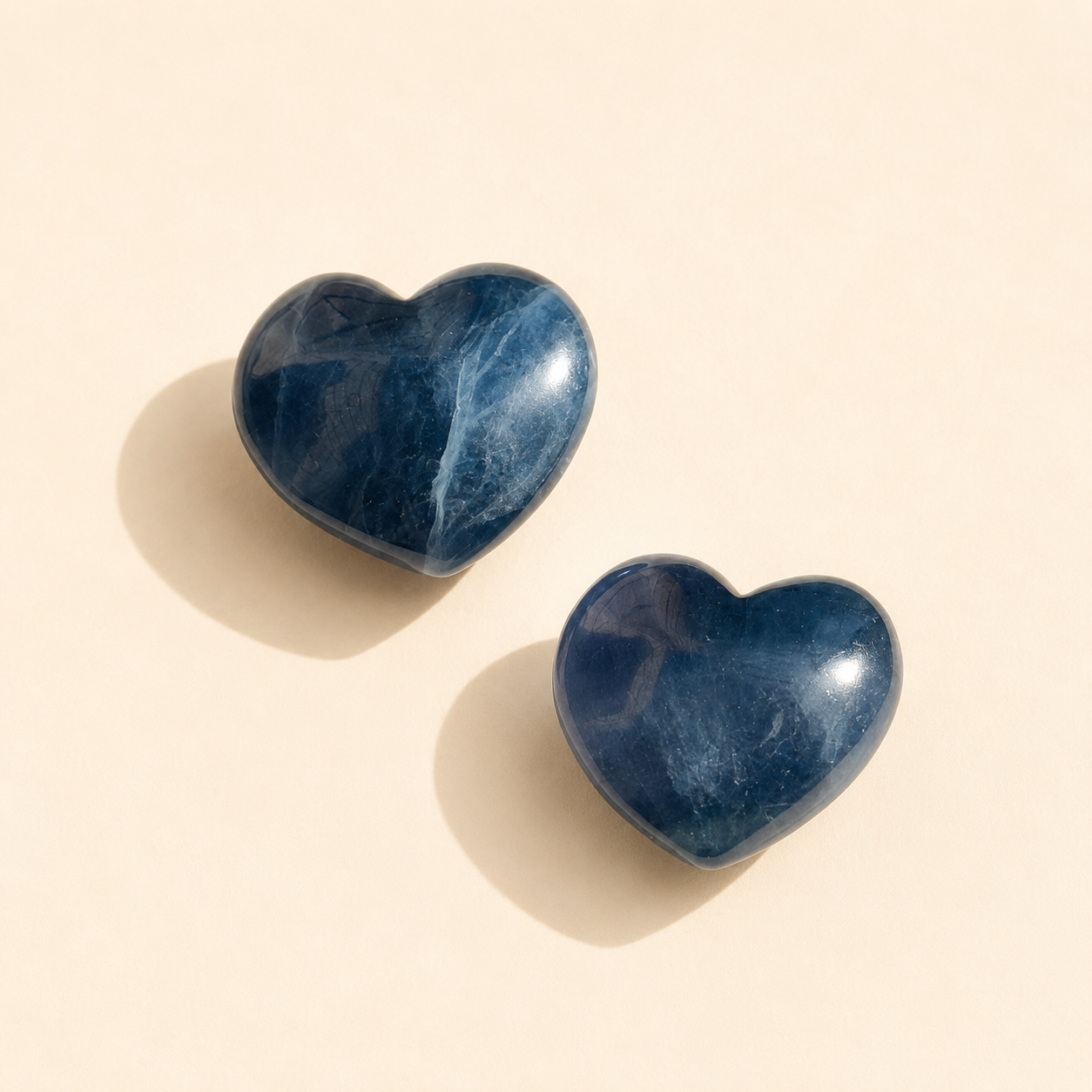 Two heart-shaped blue fluorite stone with a polished, glossy finish, displayed on a warm beige background with soft shadows.
