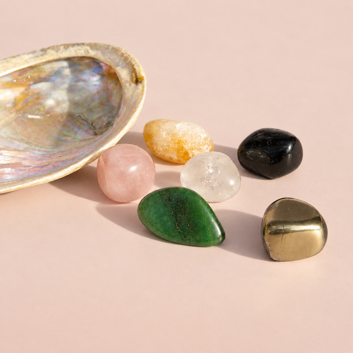 A set of seven polished tumbled gemstones in various colors arranged next to an abalone shell on a soft pink background. The stones include Pyrite, Black Tourmaline, Clear Quartz, Citrine, Rose Quartz, and Green Aventurine.