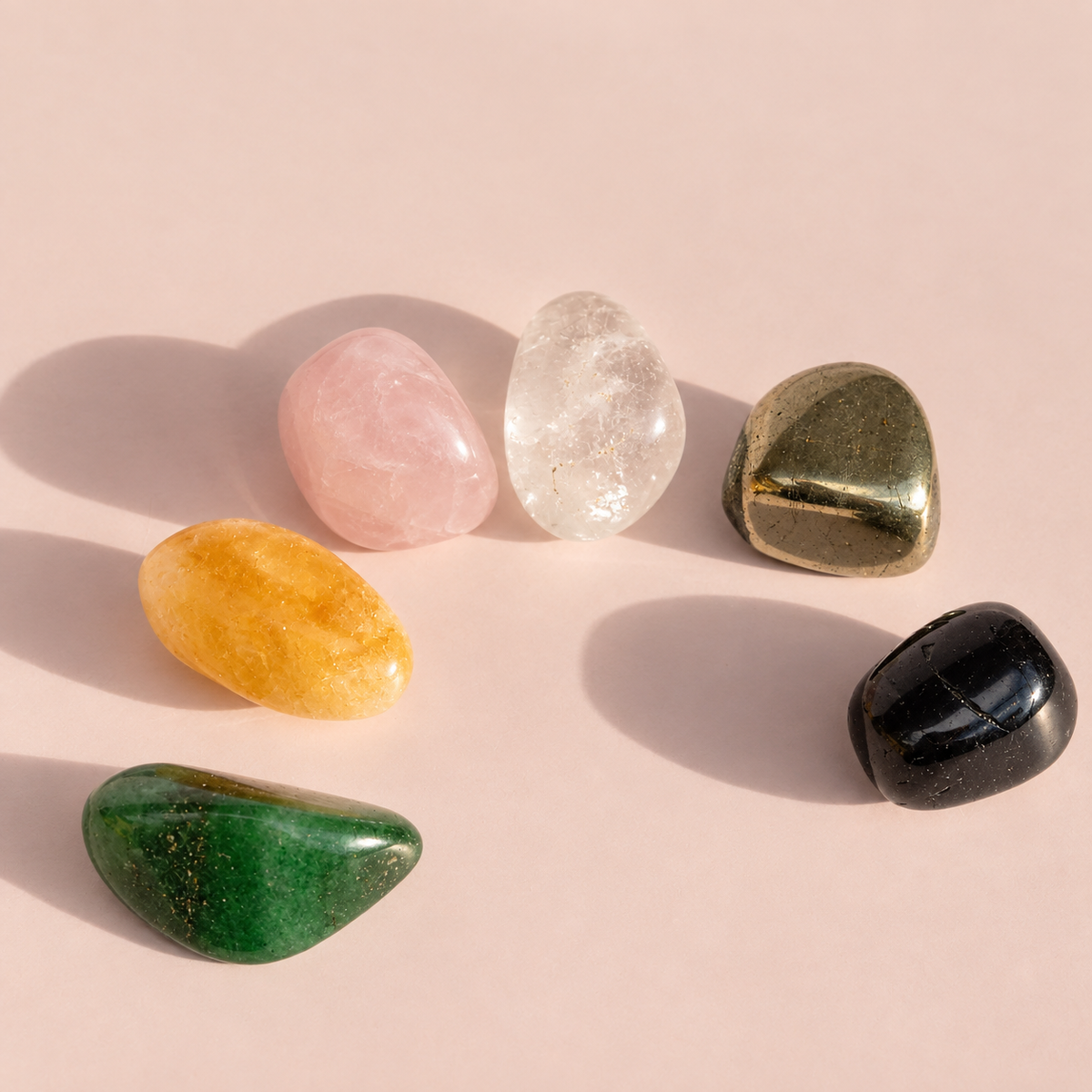 Six polished tumbled crystals in Pyrite, Black Tourmaline, Clear Quartz, Citrine, Rose Quartz, and Green Aventurine arranged on a soft pink background with shadows.