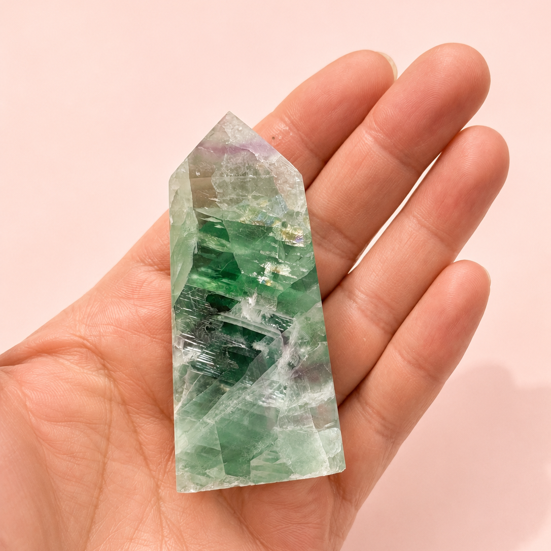 A hand holding a pointed green fluorite crystal tower against a soft pink background.