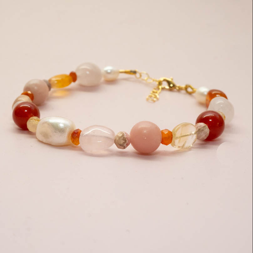 A beaded bracelet featuring a mix of polished red, translucent pink, white, and orange beads with a gold clasp, displayed on a soft pink background.