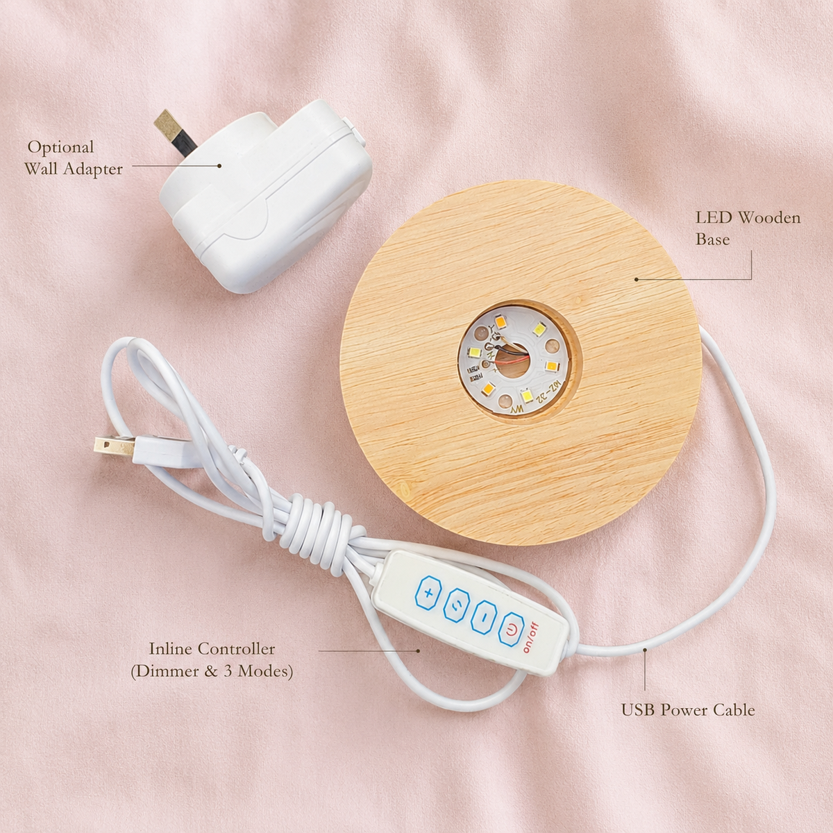 A round LED wooden base lamp with a light natural wood finish, shown with its white USB power cable, inline controller, and an optional white wall adapter on a soft pink fabric background. Text labels identify the components.