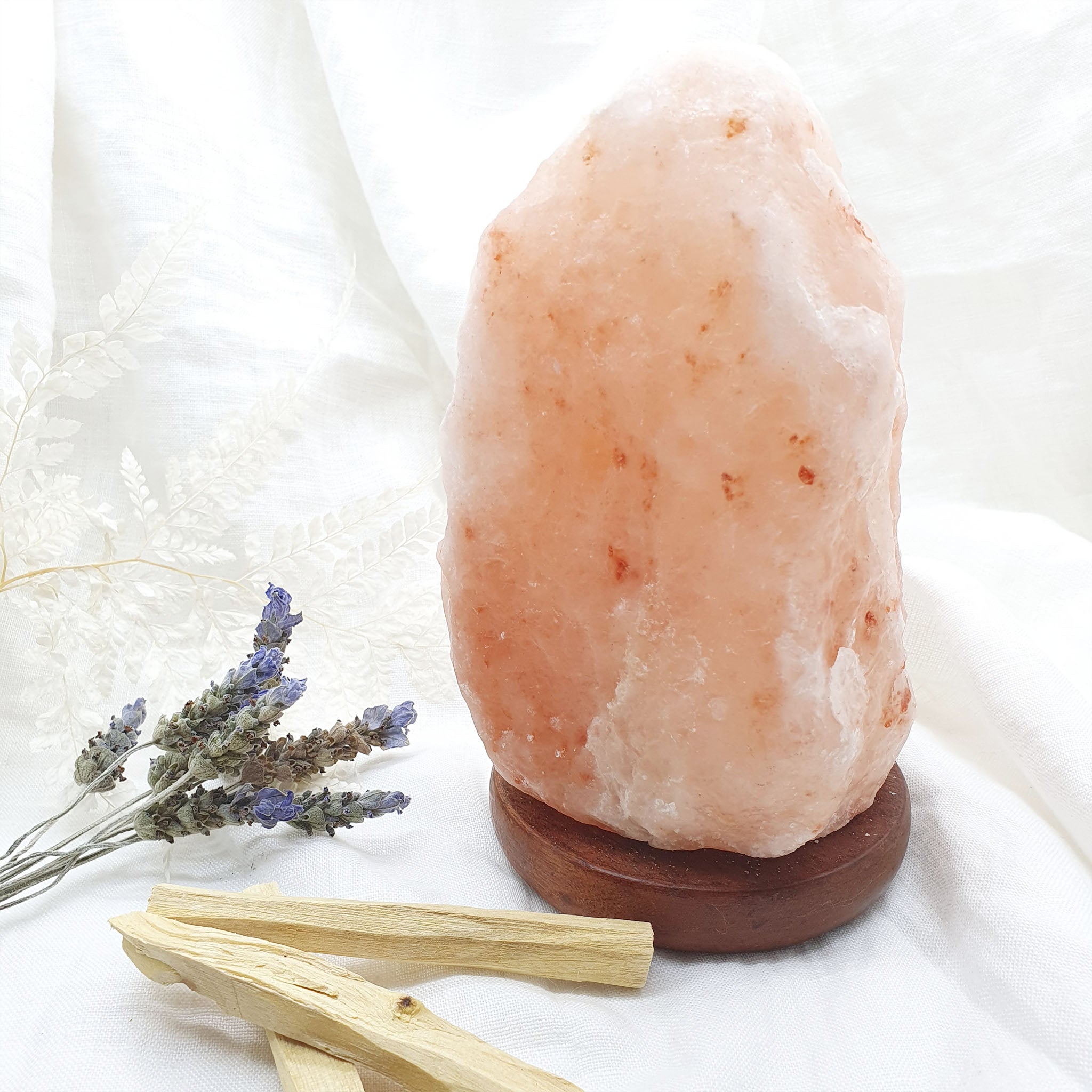 A natural Himalayan salt lamp on a wooden base with two Palo Santo sticks and some lavender sprigs in the foreground, all placed on a white surface.