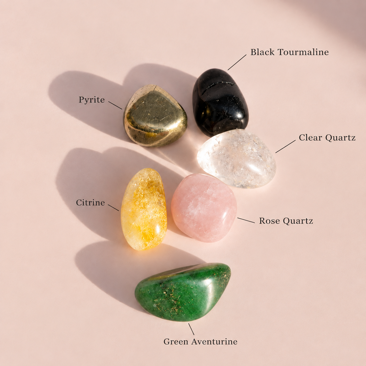 Five polished tumbled stones arranged on a pale pink background with handwritten labels identifying each stone: Pyrite, Black Tourmaline, Clear Quartz, Citrine, Rose Quartz, and Green Aventurine.