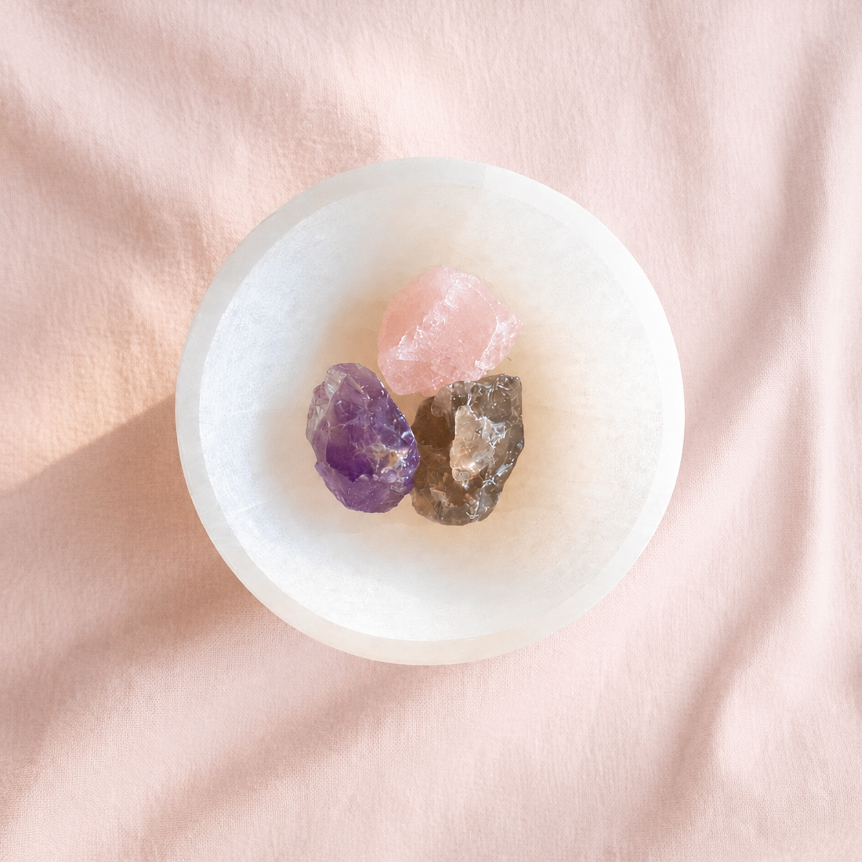 Three raw crystals inside a small selenite  crystal bowl on a soft pink fabric background.