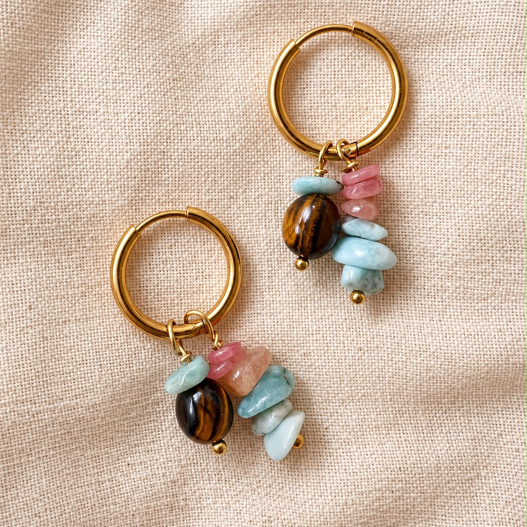 A pair of gold hoop earrings with dangling charms made of pink and light blue stone chips, a polished brown tiger's eye bead, and a light blue stone chip, displayed on a textured beige fabric background.