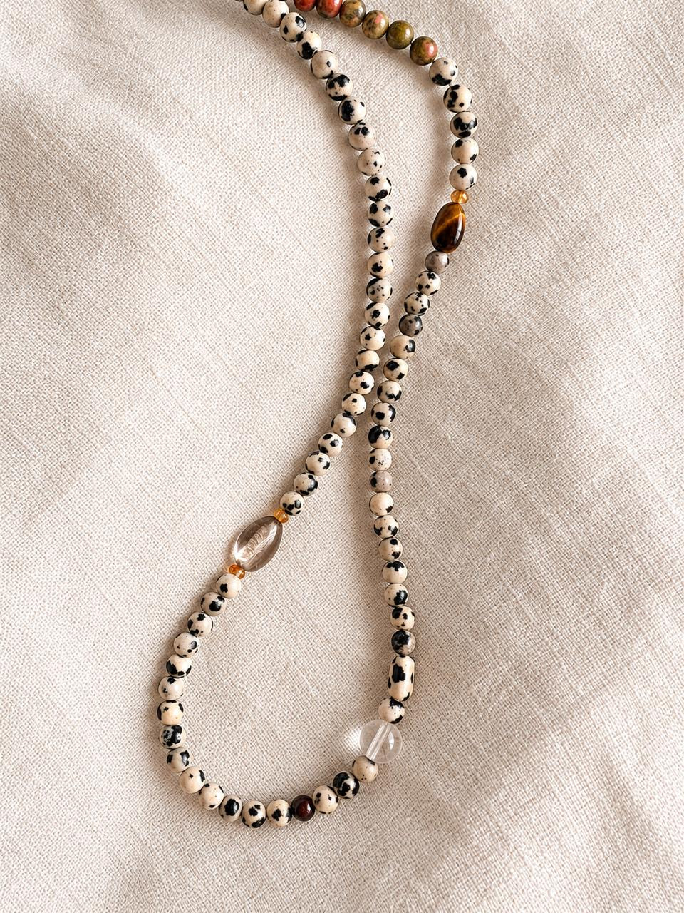 A beaded necklace with white, black, and brown patterned Dalmatian Jasper beads, a large polished brown tiger's eye bead, and a clear crystal accent, laid out on a textured beige linen fabric.