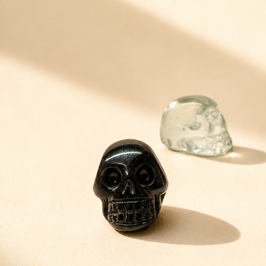A polished black tourmaline skull-shaped crystal displayed on a warm beige surface with soft shadows, alongside a translucent blue obsidian crystal skull.