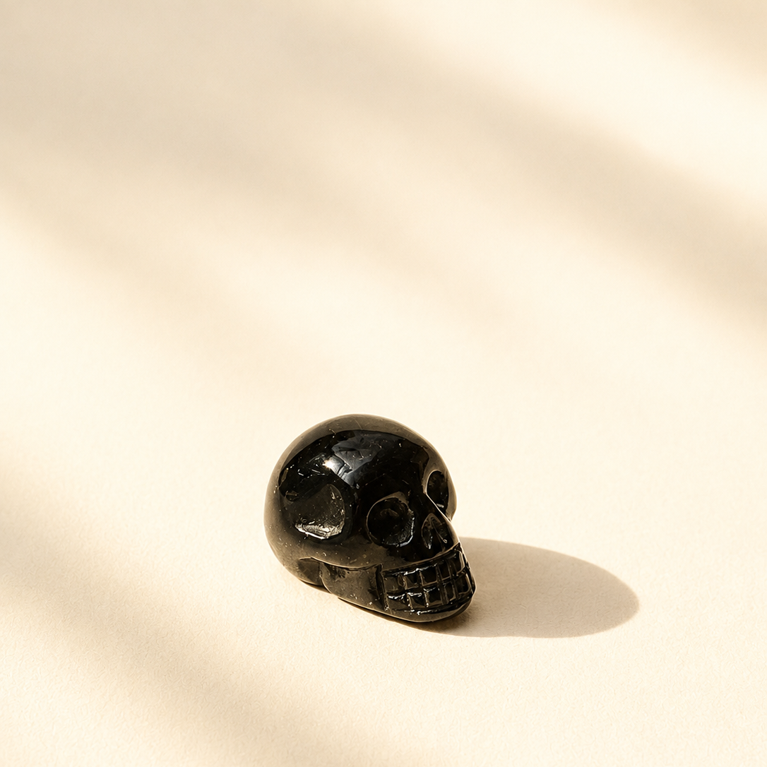 A small polished black tourmaline crystal skull figurine resting on a warm cream surface with soft diagonal shadows cast across it.