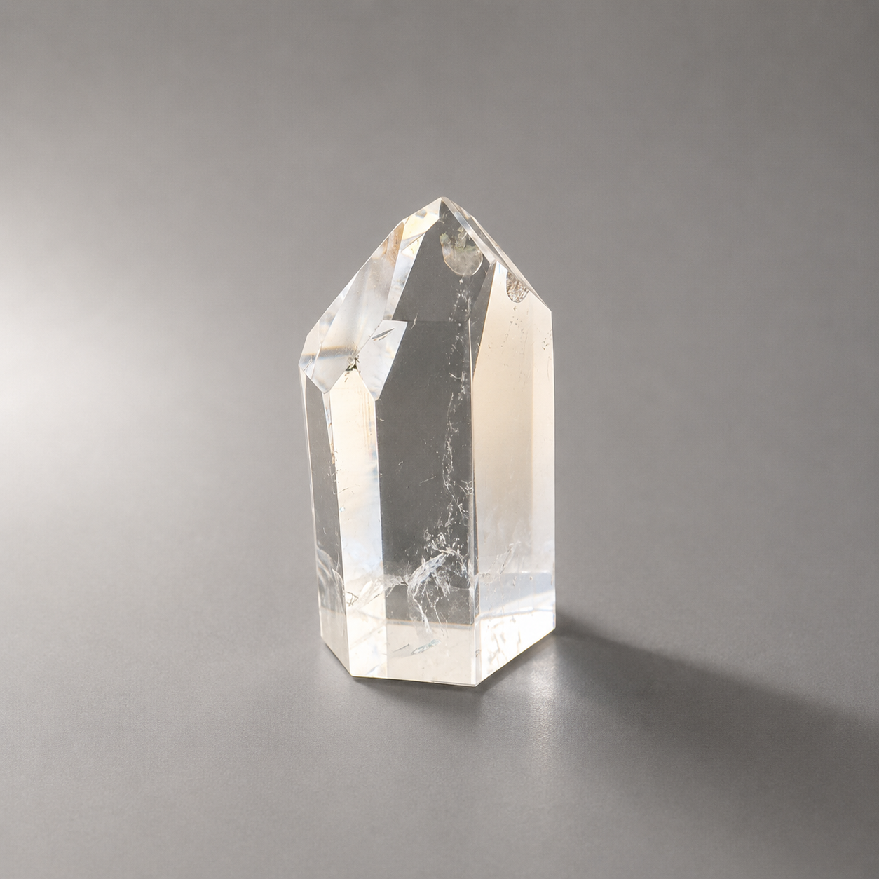 A clear quartz crystal point tower with natural inclusions, standing on a neutral gray surface against a gray background.