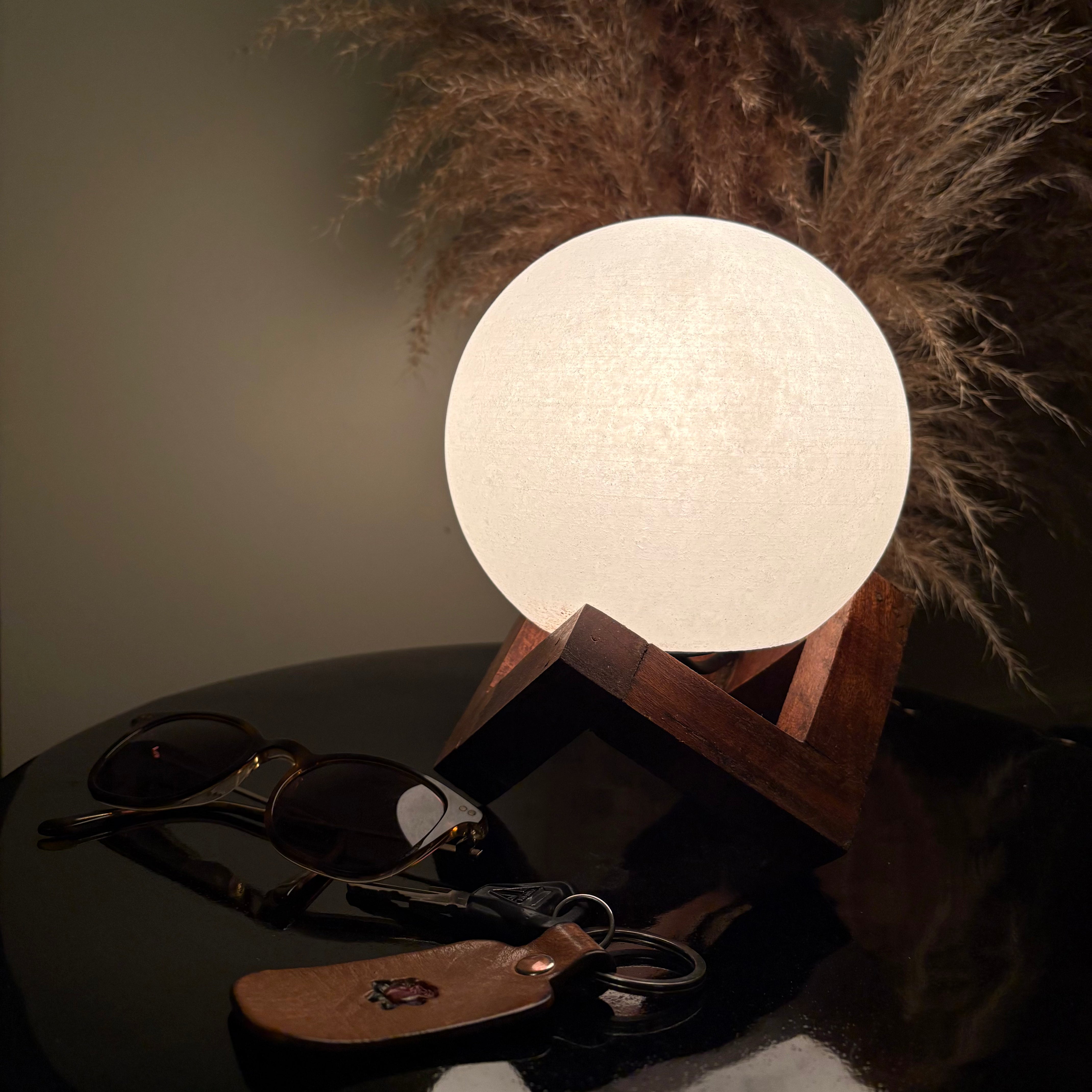 White Salt Moon lamp on a wooden stand with sunglasses and a wallet on a dark surface, pampas grass in the background.