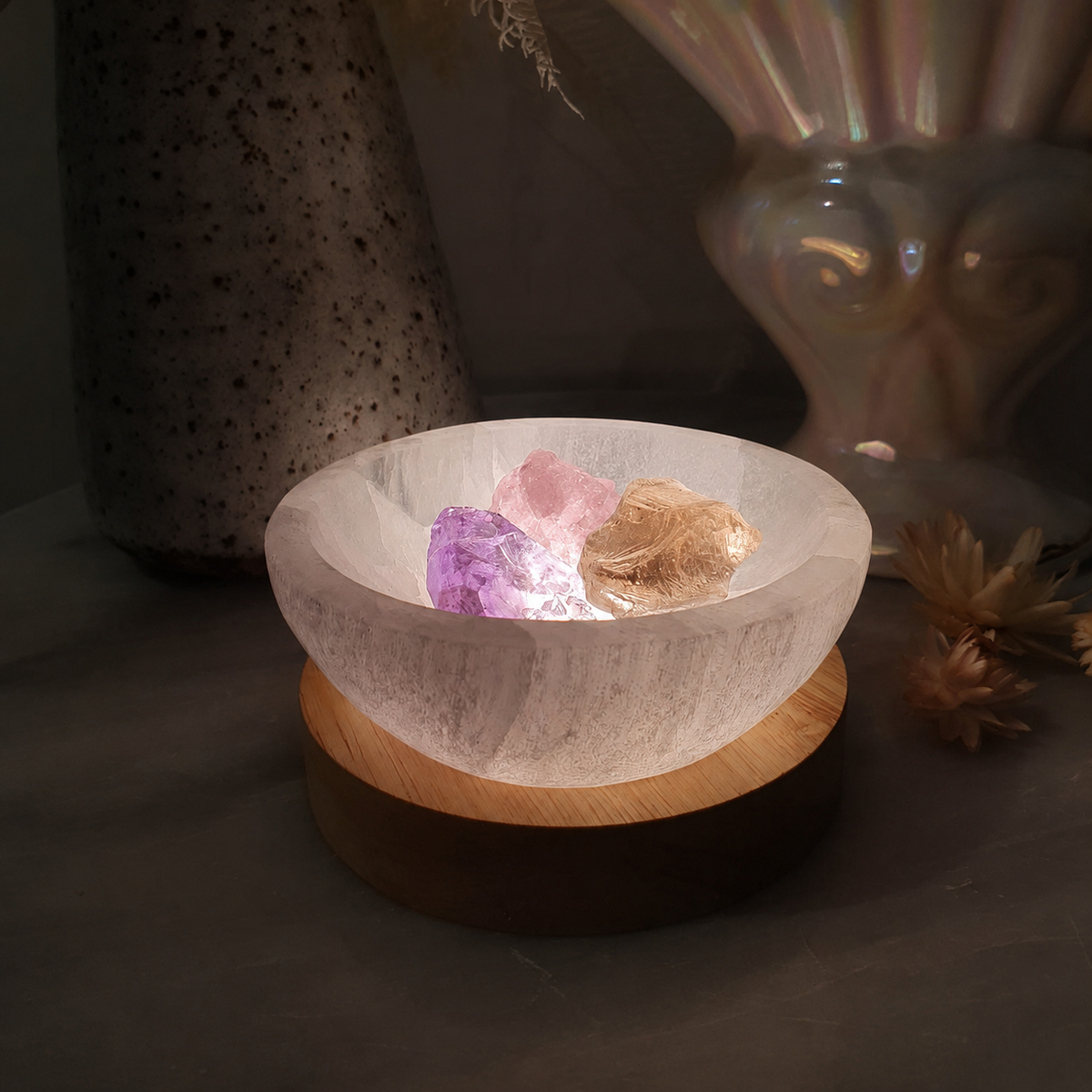 A selenite crystal bowl resting on a round wooden base, filled with rose quartz, amethyst and smoky quartz crystals, set against a dark, softly blurred background with decorative vases.