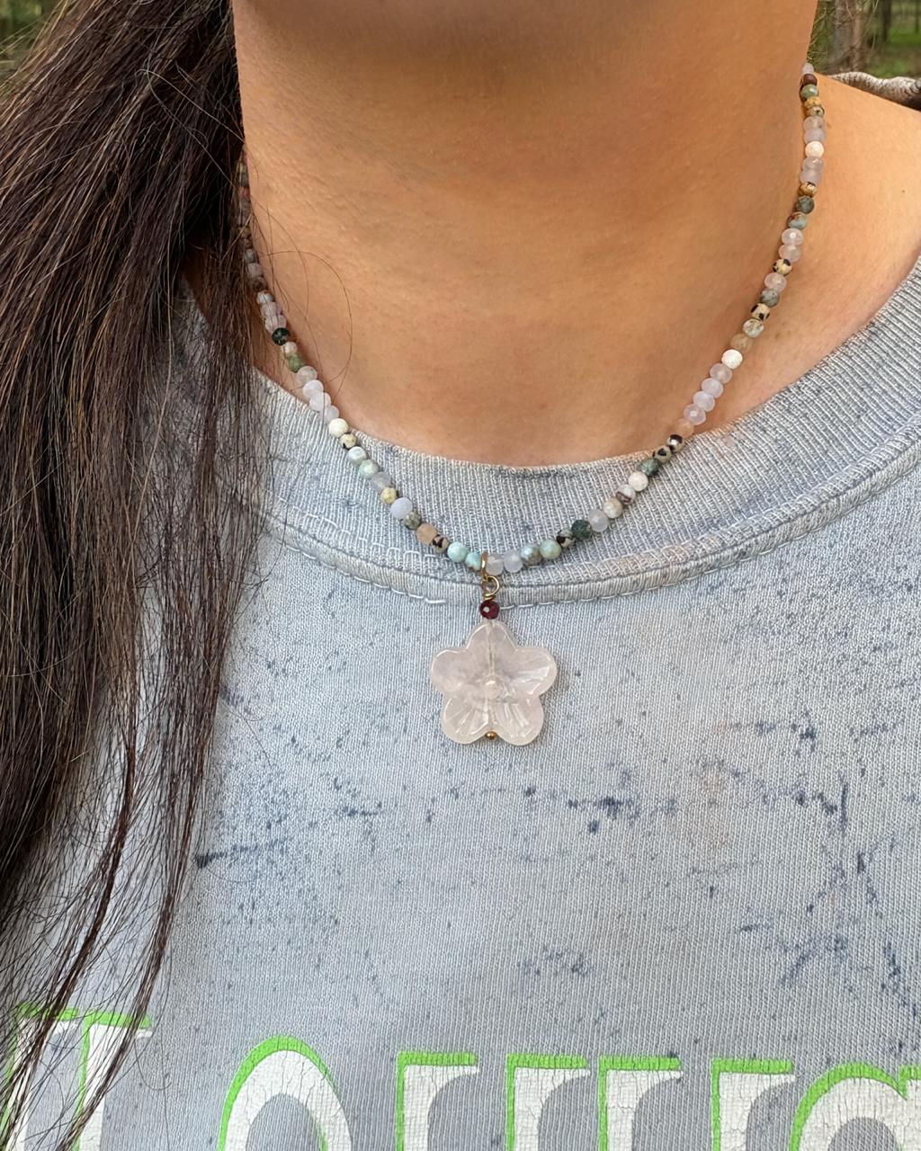 A close-up of a person wearing a beaded necklace with a pale pink flower-shaped pendant, paired with a blue sweatshirt.