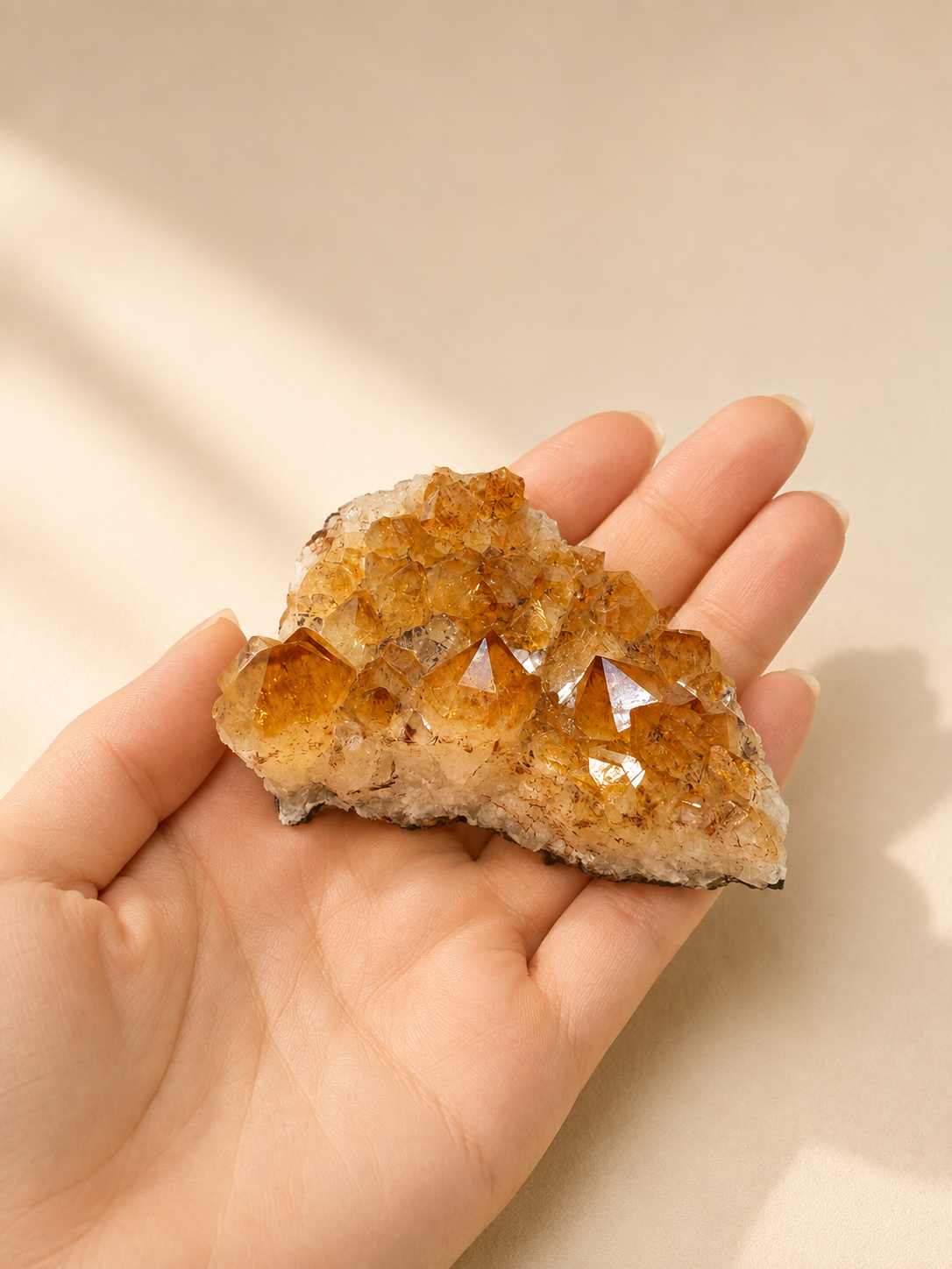 A hand holding a citrine crystal cluster with multiple pointed orange-brown and translucent crystal points on a beige background.