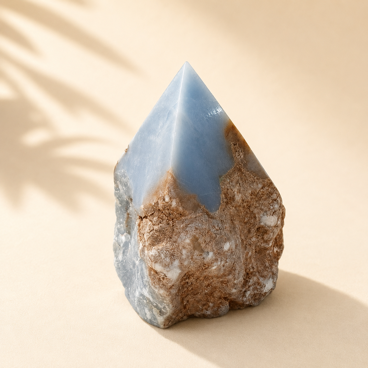 A natural pointed crystal tower with a smooth light blue top and a textured brown and white matrix base, photographed on a warm beige surface with soft leaf shadows.