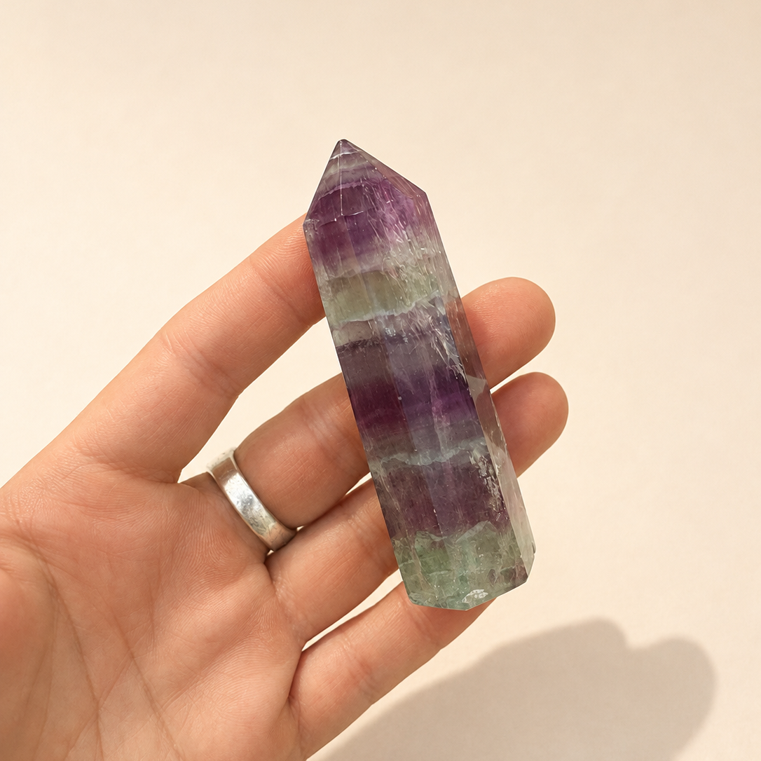 A hand holding a pointed fluorite crystal tower with visible purple, green, and white banding, against a plain beige background.