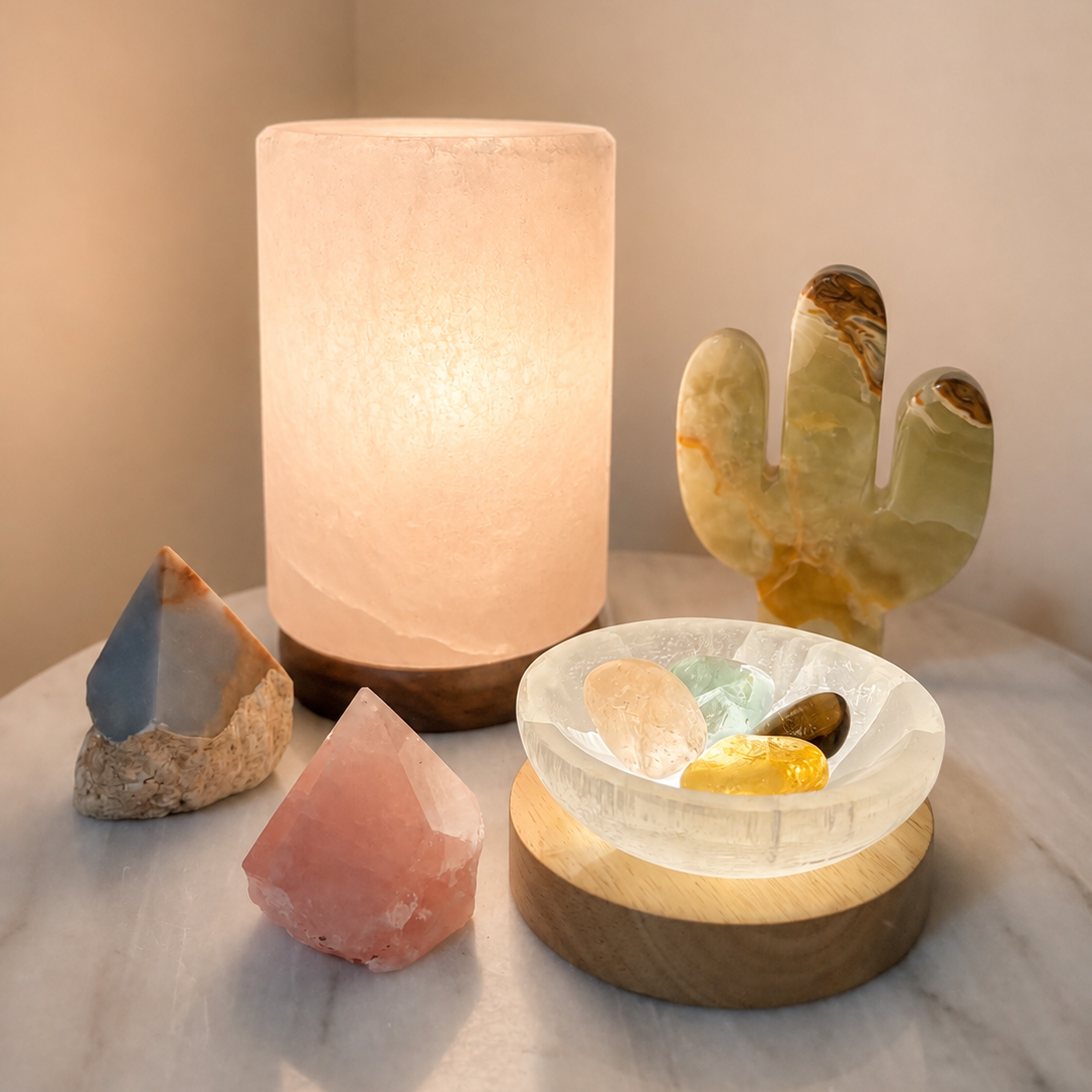 A glowing salt lamp stands on a marble surface beside several polished crystal points and a carved stone cactus, with a small clear crystal dish resting on a wooden base.