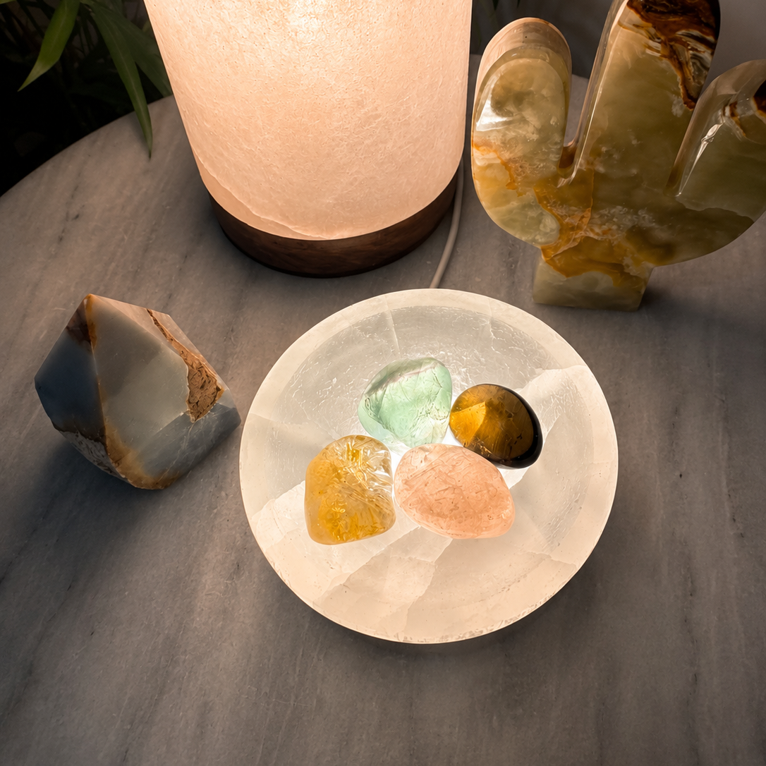 A glowing white salt lamp stands on a wooden base next to a white circular tray holding four polished crystals in green, yellow, pink, and brown. A carved stone cactus and a rough stone are also visible on a gray tabletop in a dimly lit setting.