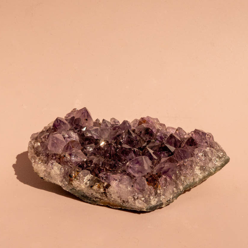 A natural amethyst crystal cluster with purple and lavender quartz points on a rough, earthy base, photographed on a solid warm beige background.