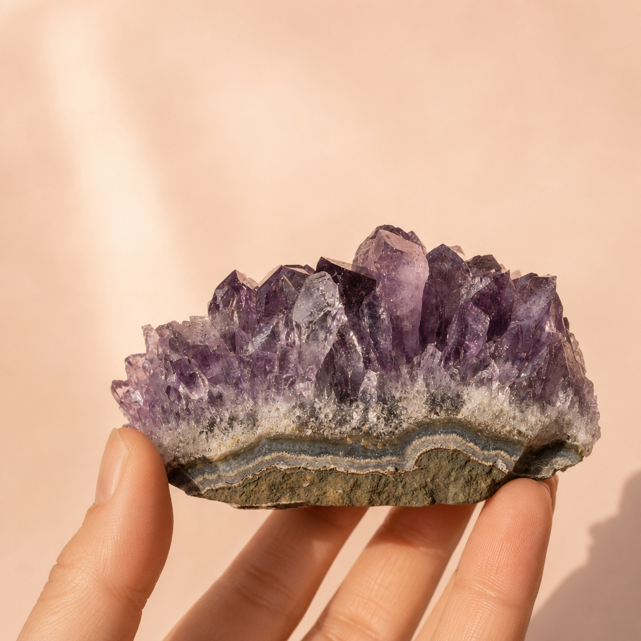 A hand holding a raw amethyst crystal cluster with a natural matrix, showing purple crystal points and a gray-brown base, set against a soft beige background.