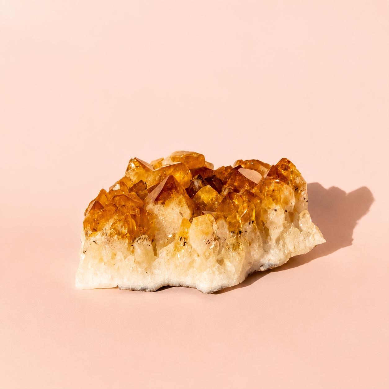 A natural crystal cluster with translucent amber-orange points on a white matrix, photographed on a solid pale pink background with a soft shadow.