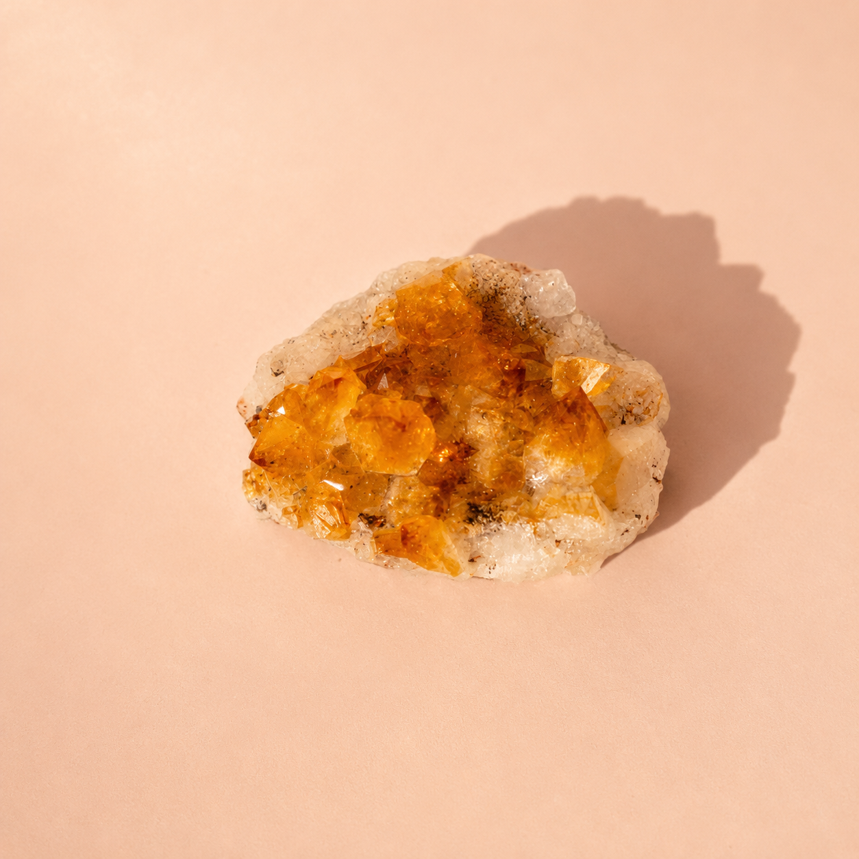 A raw citrine crystal cluster with prominent orange and yellow fluorite points on a white matrix, photographed on a solid peach-beige background with a soft shadow.