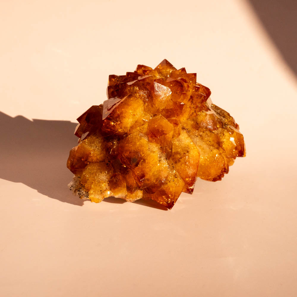 A citrine crystal cluster, photographed on a warm beige surface under direct sunlight with a soft shadow.