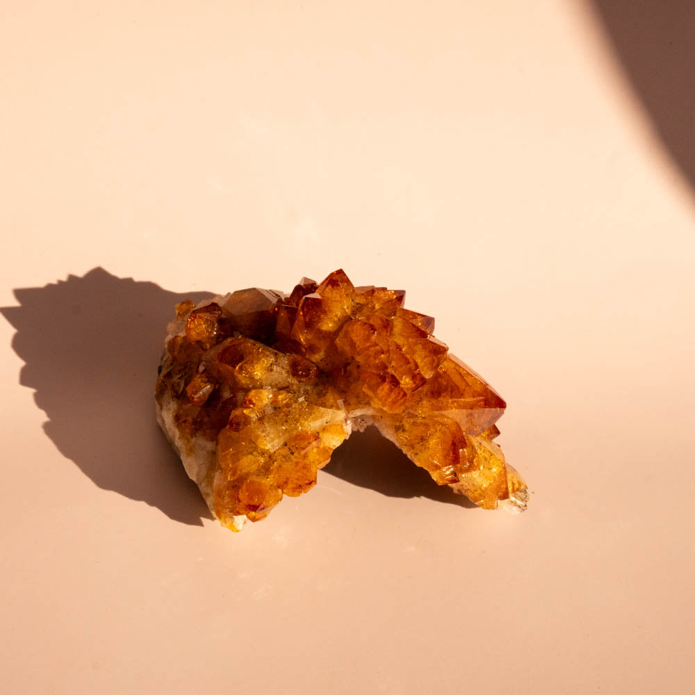 A citrine crystal cluster with sharp, textured points, resting on a warm beige surface under direct sunlight that casts a hard shadow.