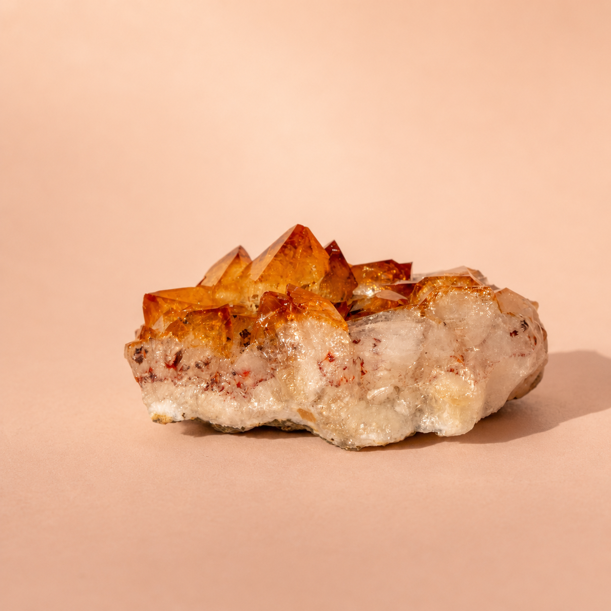 A Citrine crystal cluster featuring translucent orange-brown points rising from a rough, milky white base, photographed on a warm peach background.