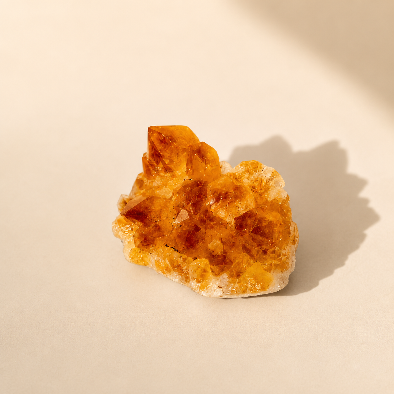 A small natural citrine crystal cluster with translucent orange and amber facets on a light beige surface, photographed in warm directional sunlight that casts a soft shadow to the right.