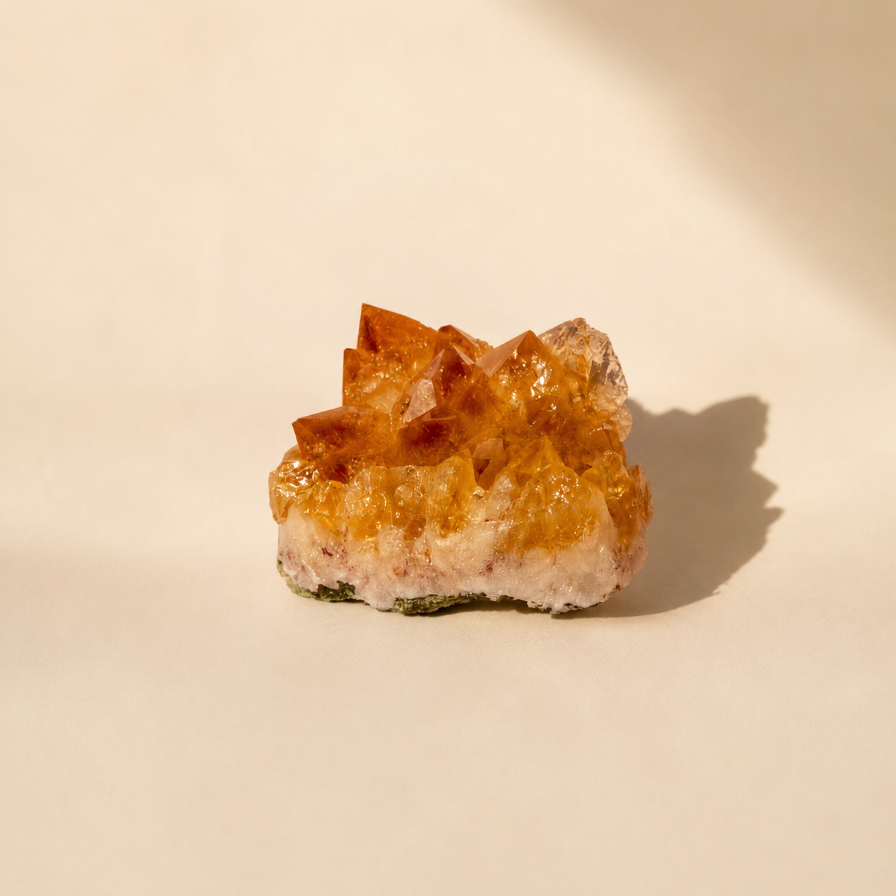 A small natural citrine crystal cluster with orange-brown translucent points and a pale pinkish base, photographed on a warm cream surface with a soft shadow to the right.