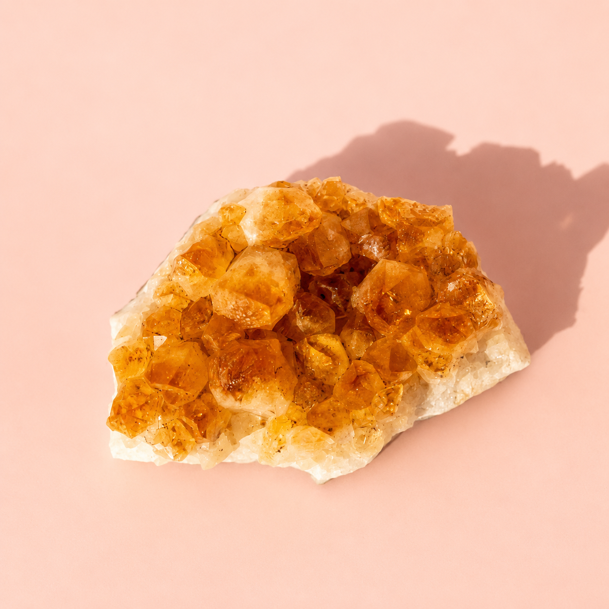 A raw crystal cluster with translucent orange and amber gemstones on a white matrix, photographed on a solid pink background with a soft shadow.