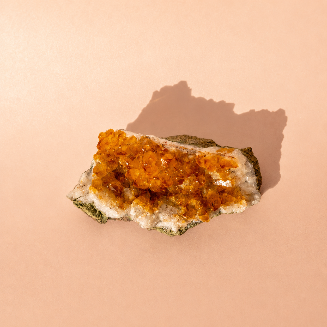 A raw citrine crystal cluster specimen with bright orange and amber druzy formations on a white matrix, set against a solid peach background with a soft shadow.