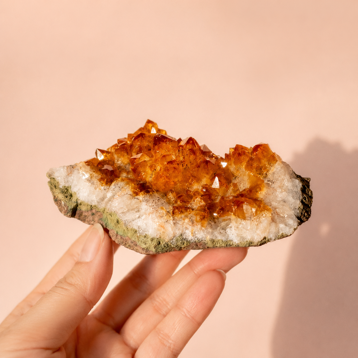 A hand holding a raw citrine crystal cluster geode specimen with prominent orange-brown crystals on a white and green matrix, set against a solid peach-pink background.