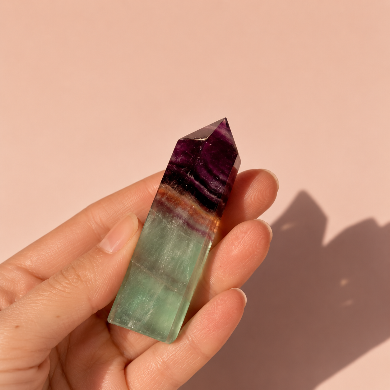 A hand holding a polished fluorite crystal tower point with distinct bands of deep purple, black, brown, light green, and milky white, set against a solid dusty pink background.