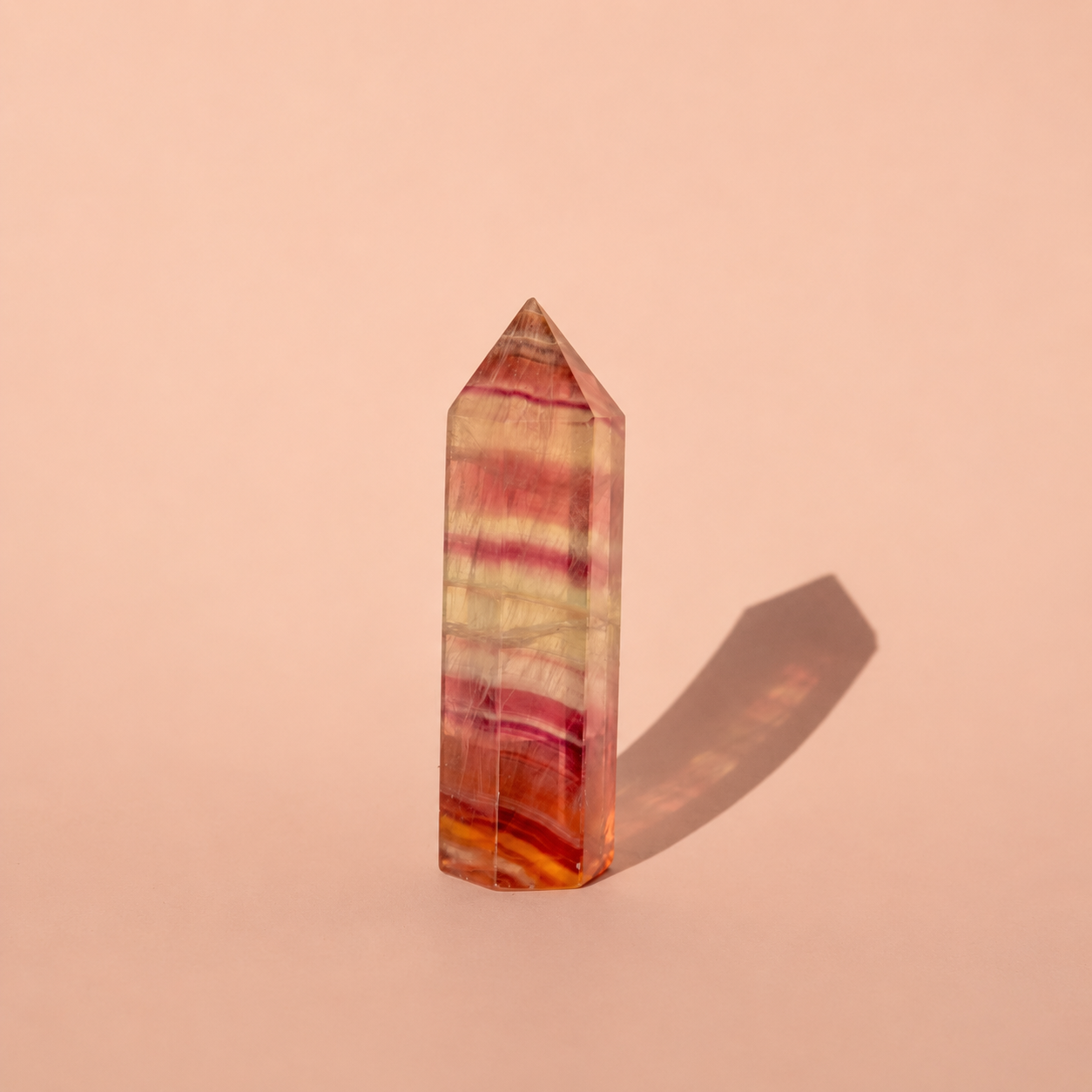 A faceted crystal fluorite tower point with pink, orange, and yellow banded stone patterns, standing upright on a solid peach-pink background with a soft shadow cast to the right.