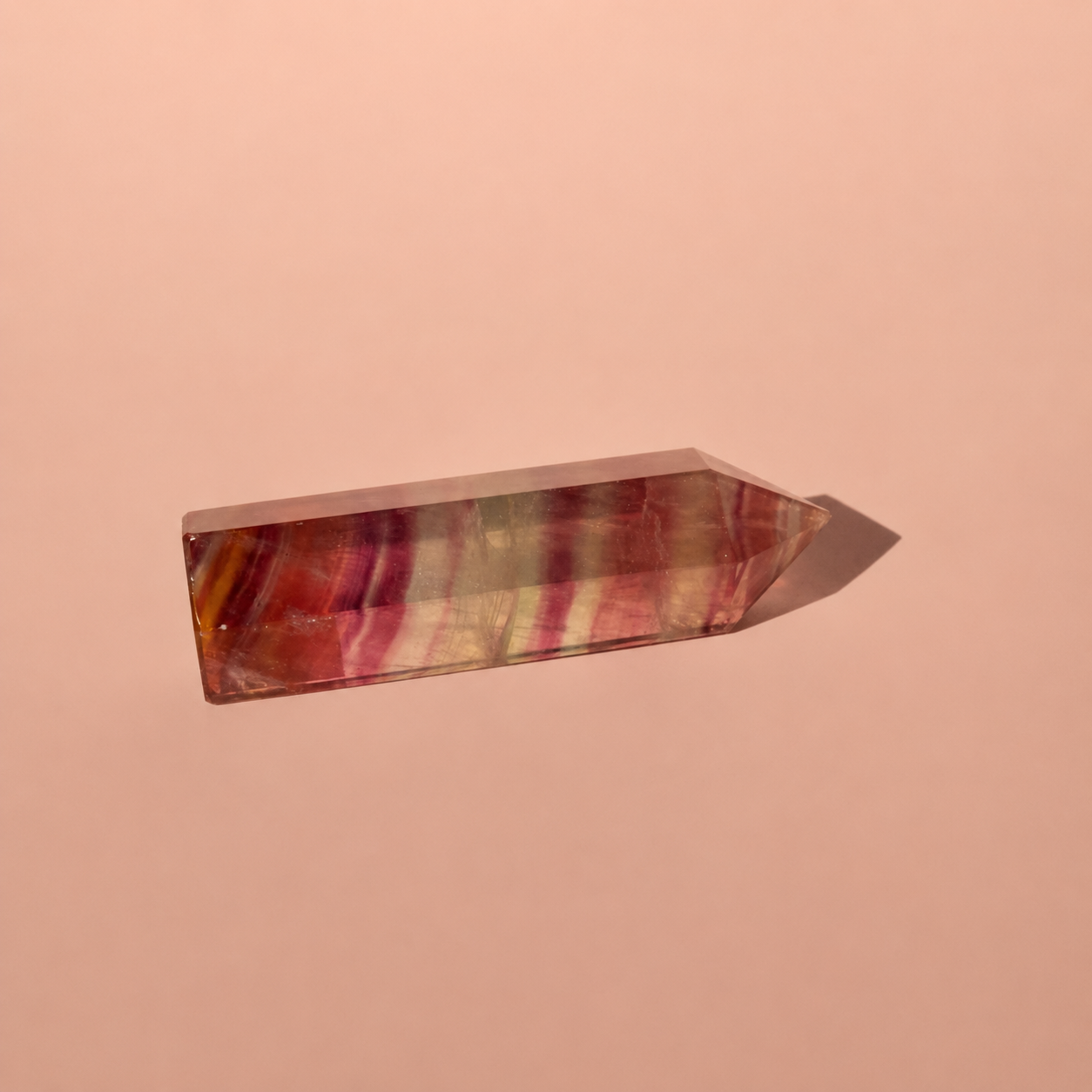 A polished pointed crystal fluorite point with pink, red, and yellow striped patterns, resting on a solid peach-pink background.