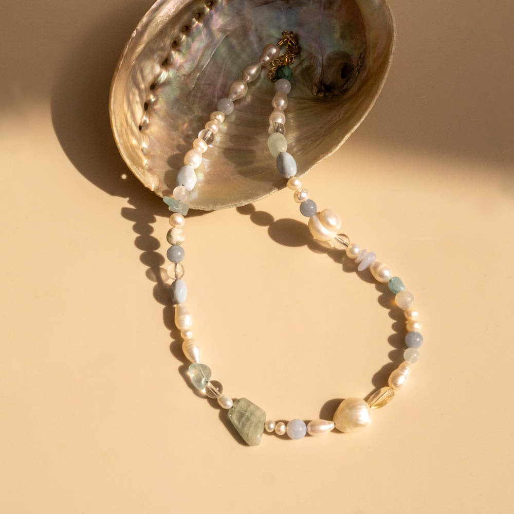 Pearl and gemstone necklace draped over a shell on a beige background