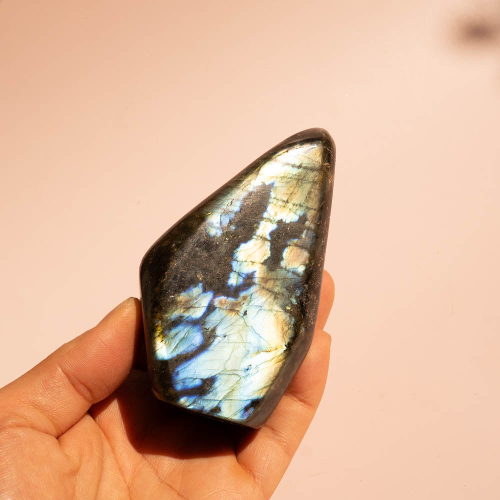 A hand holding a polished labradorite stone with a dark gray base and vibrant blue, gold, and white flashes of light, set against a soft, solid peach-pink background.