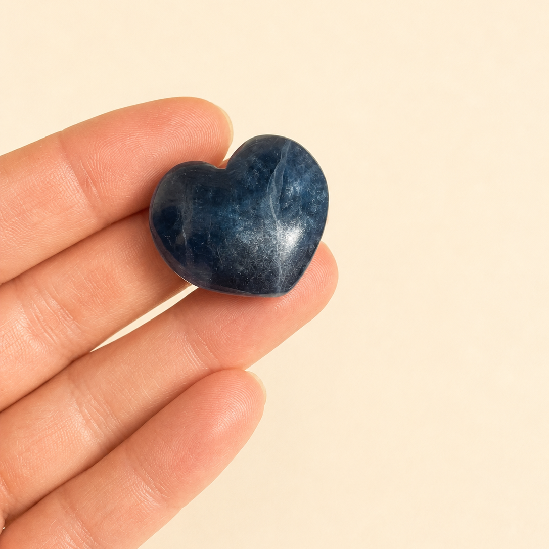 A small polished  blue fluorite crystal heart-shaped stone held on a person's fingers against a plain warm beige background.