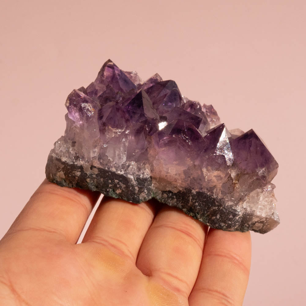 Amethyst crystal cluster held in a hand against a pink background